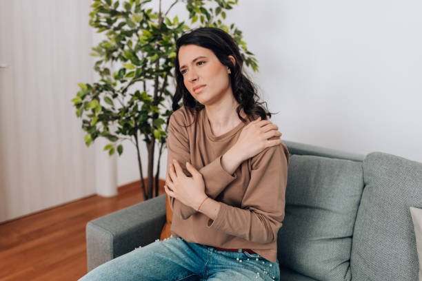 Poor young beautiful Caucasian woman feeling pain in shoulder while sitting on sofa at home