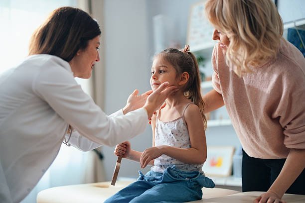 A pediatrician examines a young girl sitting on a medical bench, with her supportive mother nearby, in a bright and friendly clinic environment.